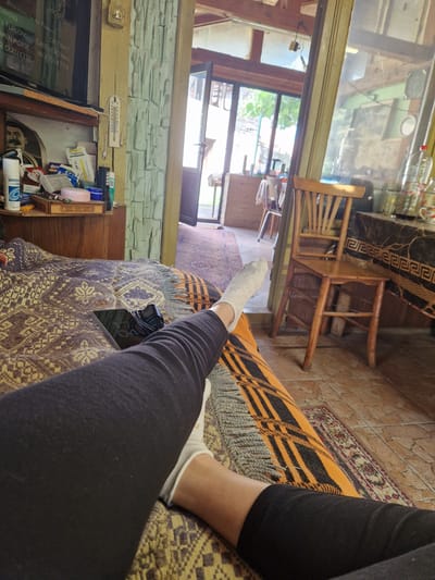 The image depicts an indoor scene, likely a room in a residence in Raduvene, Bulgaria, with an open doorway leading to an outdoor area. In the foreground, a person's legs, clad in black leggings and white socks, are visible resting on a patterned blanket. A smartphone, displaying the time 11:04, is placed on the blanket near the person's legs, along with a black electrical charger.
The room features a wooden cabinet on the left, holding various items including bottles, boxes, and a thermometer. Above the cabinet, a television screen displays text, possibly credits from a film. A rough, stone-like textured wall is visible behind the cabinet.
The open doorway reveals a sunlit outdoor space with trees and greenery. Inside the room, a wooden chair is positioned near a table covered with a dark fabric featuring a patterned border. The flooring appears to be tiled. The overall impression is one of a relaxed, domestic setting.