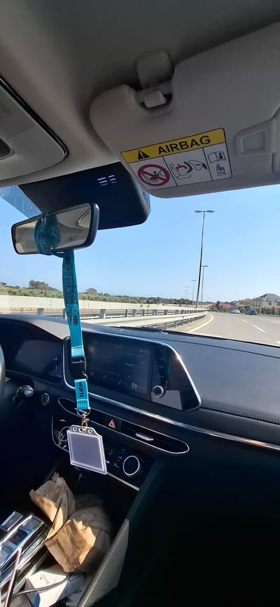 The image is taken from inside a car, looking forward and slightly to the right. The scene appears to be a daytime drive on a clear, sunny day. The sun is bright, casting reflections on the car's dashboard and creating a well-lit environment.
Prominently visible in the foreground is the sun visor with an "AIRBAG" warning label. The label has several warning symbols, including one indicating to avoid placing children in the front seat and another depicting a person being struck by an airbag. Adjacent to the visor is the car's rearview mirror, from which a turquoise lanyard hangs. The lanyard has a pattern and also displays the text "WUF13".
The car's dashboard is modern, with a large infotainment screen displaying navigation information and a circular knob for controls. Below the screen, there are climate control buttons and vents. Further to the right, a few items are placed in a compartment, including a crumpled brown paper bag and a pack of Winston cigarettes.
The view through the car's windshield shows a road curving to the right, bordered by a concrete barrier and vegetation. In the distance, there are streetlights and some buildings, suggesting an urban or semi-urban environment. The sky is a clear blue. The image is oriented vertically, with the road and landscape appearing to run upwards. The overall impression is that of a typical car journey on a pleasant day in Khatay Raion, Azerbaijan.