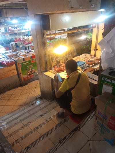 A person in a yellow shirt and dark pants sits on a red cushion, looking at a smartphone. The setting appears to be a market stall with meat displayed. Other food items are visible in the background. The stall has a light source above, and a scale and other tools are on the counter. The person is positioned on the stairs leading up to the stall. Boxes are stacked to the side of the stairs.