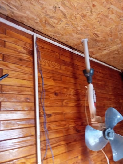 The image shows a ceiling fan mounted on a wooden paneled wall and a plywood ceiling. The fan is silver with three blades and a white motor housing. A white pipe extends from the ceiling to the fan. Several purple wires hang from the wall. The room appears to be unfinished or rustic. The image was taken in Piskivka, Ukraine.