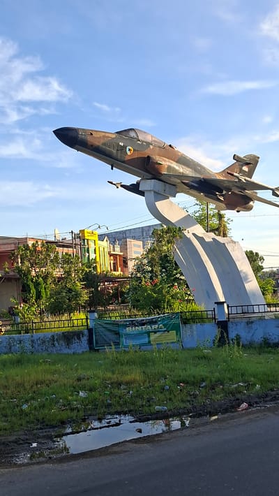 A grounded fighter jet monument, painted in camouflage brown and green, is displayed on a white, abstract pedestal against a bright blue sky with scattered white clouds. The jet is angled upwards and to the left, with its nose pointing towards the upper left corner of the frame. The cockpit is visible, as are markings on the fuselage, including a roundel with a red, black, and yellow emblem and what appears to be a Indonesian flag on the tail.
Below the monument, a black metal fence separates the display area from a patch of overgrown grass littered with various debris, including plastic bottles and wrappers. A paved road runs along the bottom of the image, with a muddy puddle reflecting the sky and surrounding environment. In the background, behind the fence and trees, are several buildings, including a prominent structure painted in bright yellow with green accents. One of the buildings has a satellite dish on its roof. The overall scene appears to be in a public area, possibly a town square or park, in Reuhattuha, Indonesia. The lighting suggests it is either early morning or late afternoon, with the sun casting long shadows and illuminating the sky.