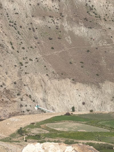A panoramic, high-angle drone shot captures the rugged, mountainous terrain of Reru, India, characterized by steep, barren slopes dotted with sparse vegetation. A prominent suspension bridge spans a gorge, connecting two sides of the valley, with two figures visible at its entrance. Below the bridge, a fertile valley unfolds, featuring terraced agricultural fields with a mix of green crops and fallow land. A few scattered trees and shrubs punctuate the landscape, adding touches of greenery to the otherwise arid environment. The overall scene depicts a remote, mountainous region with evidence of human habitation and agricultural activity.