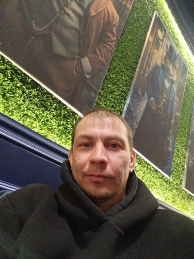 A man wearing a black coat or jacket with a high collar is posing for a selfie. Behind him is a wall decorated with artificial green turf or grass and several framed pictures. The pictures appear to depict figures in period clothing. There is also a dark blue panel behind the man. It is difficult to determine the exact location in Gus-Khrustalny, Russia without additional information.