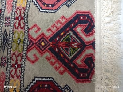 A close-up, overhead shot shows a portion of a finely woven Persian rug with a beige background and intricate geometric patterns in red, dark blue, yellow, and green. The rug features a central motif with symmetrical, stylized animal or floral shapes. To the left, a narrow, vertical band of the rug displays a repeating floral or star pattern in similar colors against a darker background. The right edge of the rug is adorned with a fringe of off-white threads. The image is watermarked in the bottom left with "REDMI 15C" and in the bottom right with "01.07.2026 01:44". The overall scene suggests an indoor setting, possibly a home or a shop in Balkanabat City, Turkmenistan, where such rugs are a traditional craft. There are no people or animals present.