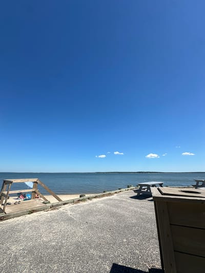 This media file depicts a clear, sunny day at the beach, likely in the Town of Riverhead, USA. The image is oriented vertically, with the horizon line running diagonally from the bottom left to the top right.
The scene is divided into two main parts: the sky and the water. The vast expanse of the sky is a brilliant, cloudless blue, with only a few wispy white clouds scattered across it, suggesting a warm and pleasant weather. Below the sky, the calm, blue water stretches out, meeting the shore. A sandy beach is visible along the water's edge.
In the foreground, on the right side of the image, is a wooden boardwalk or pier. Part of a wooden picnic table with benches is visible on the boardwalk, casting a shadow on the textured asphalt path that runs alongside it. Further along the boardwalk, towards the bottom of the frame, there is a wooden structure with a blue and red umbrella, and beach chairs, suggesting a place for relaxation and enjoyment of the beach. A few pairs of flip-flops are also visible on the wooden deck. The overall setting conveys a sense of leisure and outdoor activity. The time of day appears to be midday, given the bright sunlight and shadows.