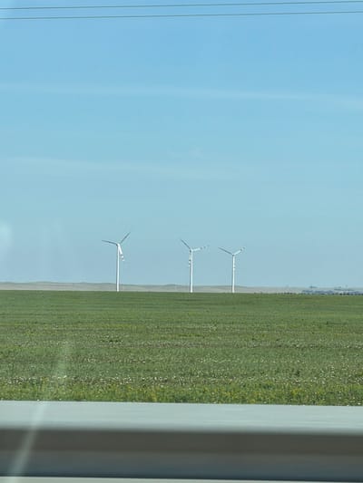 The image depicts a wide, flat landscape under a clear blue sky. In the middle ground, three white wind turbines stand in a row, spaced apart. They have three blades each, with the blades of the first turbine visible and rotating. The turbines are erected on white cylindrical towers. The foreground is dominated by a field of green grass, dotted with small white and yellow flowers, suggesting a natural or agricultural setting. A horizontal light gray band runs across the bottom of the image, possibly a road or barrier, with some blurry reflections. In the far background, a low, hazy horizon line is visible, with what appear to be distant structures or a body of water. The scene is devoid of human presence. The overall impression is one of open, rural space with a focus on renewable energy infrastructure. This landscape is consistent with the flat terrain often found in the Turgay region of Kazakhstan, known for its wind farms.