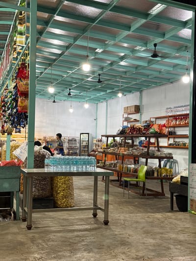 The image depicts the interior of a store located in Grong-Grong, Indonesia. The store appears to be a small, local market or convenience store, with shelves stocked with various food items, snacks, and beverages. In the foreground, a metal table holds rows of bottled water. To the left, large bags of what appear to be snacks are visible. In the background, a person wearing a blue and white patterned shirt is standing near shelves filled with packaged goods. Another person is partially visible to the left of the main figure, facing away from the camera. The store features a teal-colored metal frame supporting the roof, which is covered with insulation panels. Ceiling fans are mounted on the roof. The lighting is provided by overhead bulbs. The floor is made of concrete. The store's exterior is not visible, but the interior suggests it is a functional retail space.