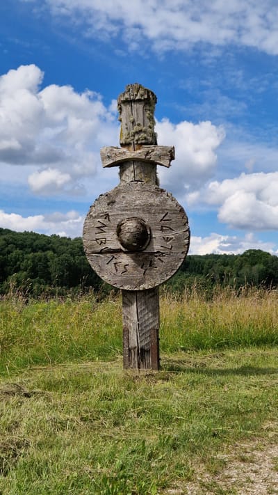 This media file features a wooden cross adorned with carvings, standing in a grassy field under a bright blue sky with fluffy white clouds. The cross is made of rough-hewn wood, weathered and covered in patches of lichen. A circular wooden disc is attached to the vertical beam of the cross, and this disc also bears carved symbols.

Visible text on the wooden disc appears to be in a runic script. One segment of the inscription is clearly legible and reads: "MBVTS". Below this, another partially visible inscription seems to say "NLAGN". The overall scene suggests an outdoor location, possibly in a rural or historical setting. The lighting indicates it is daytime, with the sun casting shadows. The sky is clear and sunny, suggesting fair weather. The background shows a dense line of green trees, possibly a forest, and a sloping field of dry grass and wildflowers. The composition is vertical, with the cross dominating the center of the frame.