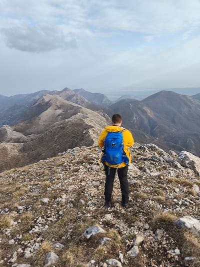 A lone individual stands on a rocky mountain ridge in Mali Mikulići, Montenegro, facing away from the camera and overlooking a panoramic mountainous landscape. The person is an adult male, viewed from behind, wearing a bright yellow jacket, dark trousers, and a blue Osprey brand hiking backpack. A hiking pole is visible near his left leg. The immediate foreground consists of uneven ground covered with light grey rocks, pebbles, and sparse patches of dry, yellowish-brown grass. Beyond, a series of barren, dark grey and brown mountain peaks and ridges extend into the hazy distance. The sky above is partly cloudy, featuring areas of light blue sky interspersed with light grey and white clouds.