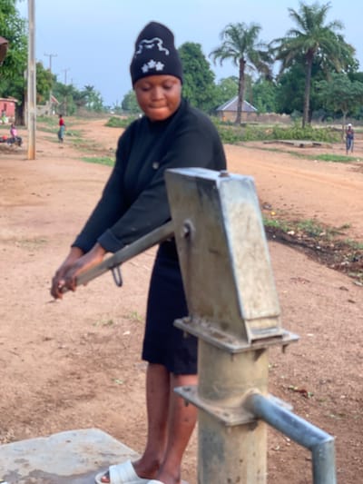 The image depicts an outdoor scene in Ningo-Sarki, Nigeria, focusing on a woman operating a hand pump. The woman, positioned prominently in the foreground, is dressed in a dark, long-sleeved top and a dark skirt or shorts. She wears a black head covering adorned with distinct white patterns, resembling a stylized bear or cloud shape alongside stars or flowers. White sandals are visible on her feet. Her hands are placed on the handle of the metal hand pump, indicating she is actively drawing water.
The hand pump itself is robust and appears well-used, made of metallic components. The immediate ground around the pump is dry, reddish-brown earth, suggesting an unpaved or rural setting. In the background, a wide dirt road stretches into the distance, bordered by verdant tropical foliage, including several tall palm trees. Further down the road, indistinct figures are visible, along with glimpses of distant structures or utility poles, indicating a settlement.
The lighting suggests it is daytime, with soft, diffused light, possibly under an overcast sky or during the gentler hours of morning or late afternoon, as there are no harsh shadows. The overall atmosphere is one of daily routine and community life, centered around a vital water source. No explicit text is clearly discernible in the image, aside from the patterns on the woman's head covering.