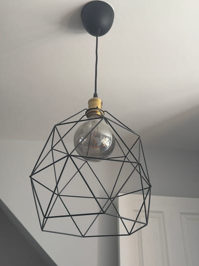 A close-up shot presents a modern wall-mounted light fixture against a neutral wall backdrop. The fixture comprises a spherical light bulb housed within a black, geometric wireframe, connected to a black, dome-shaped mount affixed to the wall via a black cable. The bulb is transparent, with visible internal elements. To the right of the fixture, a white, paneled surface is partially visible, suggesting a door or wall feature. The scene lacks people or identifiable actions, concentrating on the aesthetic of the lighting element. The photograph does not indicate a location in Guildford, United Kingdom.