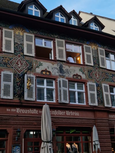 The image depicts the facade of a historic building in Rheinfelden, Switzerland, characterized by ornate painted murals and architectural details. The building features multiple windows with white shutters, some of which are open. A man in a white shirt is visible in one of the open windows. The facade is adorned with various decorative motifs, including sunburst patterns, animal figures, and intricate floral designs.

On the right side of the building, painted text reads "bis 1884 Braustätte des einfelder Urbräu," indicating the building's historical association with a brewery. A closed light-colored umbrella is tied to a pole extending from the building. A sign above a window reads "Dore".

Additional text on the building reads "Begrundet von Franz-Jos Dietschy A-D-1799" and "Renovist ven Labich-Dietschy 1896-199," providing historical context about the building's founders and renovations. The architectural style is typical of historic buildings in European cities. The image captures the charm and historical character of Rheinfelden.