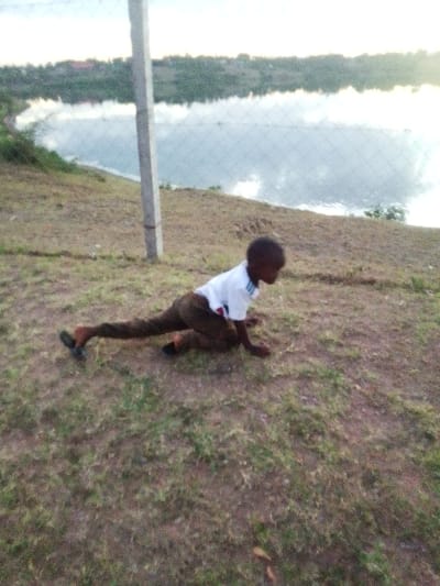 A single individual, appearing to be a young male of short stature, is depicted outdoors in a low, crawling-like posture on uneven ground. The individual wears a white short-sleeved t-shirt with blue and red accents on the collar, and dark brown or black trousers. A dark-colored sandal is visible on the left foot, and another on the right. Both hands and the right knee are on the ground, while the left leg is extended backward. The immediate ground cover consists of sparse grass and exposed soil. A light-colored vertical pole stands on the left, with a mesh fence extending horizontally behind it and the individual. Beyond the fence, a large body of water reflects the sky, with a vegetated shoreline visible in the distance under an overcast or hazy sky. The scene is located in the city of Central Kasipul ward, Kenya, adjacent to a significant body of water.