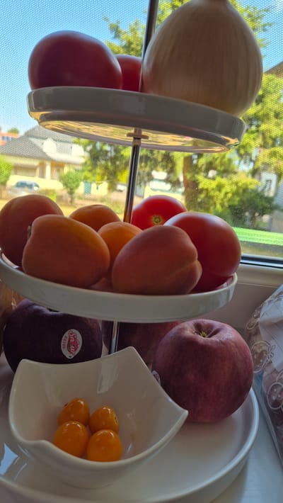In Miltenberg, Germany, a tiered fruit stand is positioned near a window, offering a view of trees and houses outside. The top tier of the stand holds two red tomatoes and a large, light-brown onion. The middle tier is laden with several apricots and two more red tomatoes. Below this, a dark red apple with a "Snow" sticker is visible, accompanied by a bowl containing four small, bright orange cherry tomatoes. Another red apple is partially visible to the right of the cherry tomatoes. The overall scene depicts a collection of fresh produce arranged on a multi-level stand.