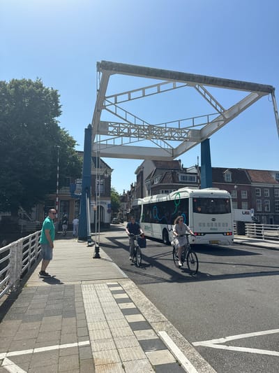 A group of people are on a bridge in Zwolle, Netherlands. The image is taken on a sunny day. On the right side of the image, a white bus is driving on the road. A woman is riding a bicycle on the road in front of the bus. A man is also on a bicycle further down the road. On the left side of the image, a large metal structure, likely part of the bridge, is visible against a clear blue sky. Below the bridge structure, a man in a green t-shirt and denim shorts stands on the sidewalk, looking out. He appears relaxed. Further down the sidewalk, a red and white barrier is down, suggesting the bridge may be in the process of opening or closing. Trees and buildings are visible in the background. The scene suggests a typical day in the city, with people commuting and enjoying the weather. The text "RR Reis" is visible on the side of the bus, and a license plate "75-BHZ-7" is also visible.