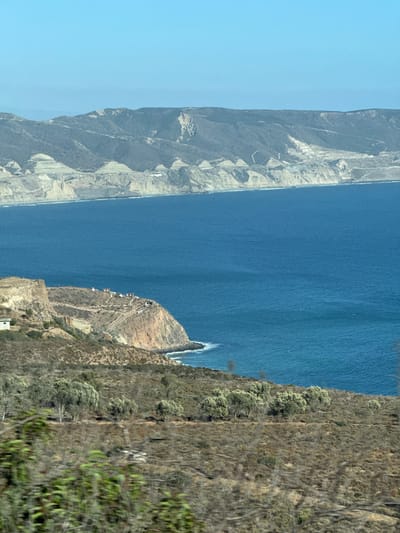 The image depicts a coastal landscape with a vast expanse of blue ocean meeting a rugged, mountainous shoreline. The distant mountains are barren and layered with pale sediment, suggesting arid conditions or extensive mining operations. In the middle ground, a prominent, dry, and rocky bluff juts into the sea, characterized by sparse vegetation and a winding path leading to a cluster of white buildings on its summit. A small, white structure is visible on the lower left slope of this bluff. The foreground features a densely vegetated hillside, with the foliage appearing dry and muted in color, interspersed with a few olive trees and shrubs. The overall impression is one of a natural, undeveloped coastal environment, possibly in a region with limited rainfall.