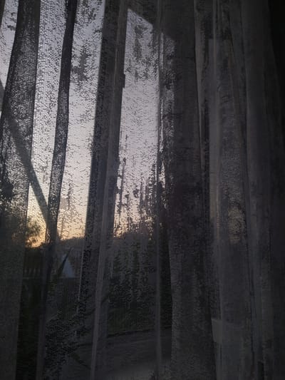 The image depicts a view through a sheer, grey curtain. The curtain is draped in vertical folds, with a fine, patterned texture visible upon close inspection. Through the semi-transparent fabric, the faint outline of a window frame is discernible on the left side, suggesting the perspective is from indoors looking out.
Beyond the curtain, the scene appears to be an outdoor environment at dusk or dawn, characterized by a soft, diffused light. Silhouetted trees and structures are visible, hinting at a residential or semi-rural setting. The sky exhibits a gradient of pale colors, transitioning from a soft orange or peach near the horizon to a muted grey or lavender higher up.
The location specified, "Мамлют қаласының әкімшілігі" (Administration of Mamlyut City), Kazakhstan, provides context for the outdoor elements. While the image itself does not contain specific landmarks or identifiable features of this administrative building or its surroundings, it can be inferred that this is a view from a window within or adjacent to that area, looking out onto the landscape as twilight sets in. There are no people or overt actions depicted in the image. The focus is on the interplay of light, shadow, and texture through the curtain, creating a mood of gentle fading daylight.