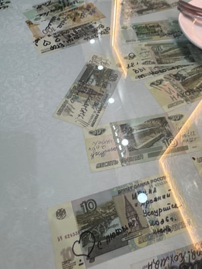 The image shows several Russian banknotes scattered on a white, patterned surface, possibly a tablecloth. The banknotes are marked with handwritten messages and drawings in black ink. The messages appear to be in Russian and include names, dates, and personal messages. Some banknotes also have drawings, such as hearts. The lighting suggests an indoor setting, with reflections from overhead lights visible on the surface. There are no people visible in the image. The banknotes are of various denominations, including 10 and 100 rubles. The surface has a subtle floral or damask pattern. The banknotes are oriented at various angles, and some are partially obscured by others. The overall impression is of a casual or informal setting where these banknotes have been left or used. There is no discernible information within the image that would indicate the location in Suifenhe, China.