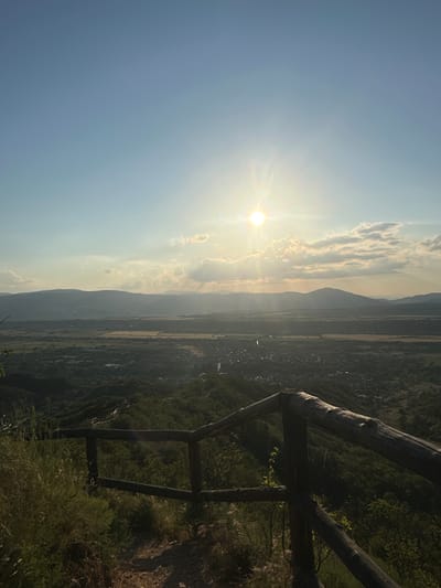 The image depicts a panoramic landscape view from an elevated position overlooking a valley in the city of Stob, Bulgaria, under a setting or rising sun. The upper portion of the frame features a clear blue sky, transitioning to lighter tones towards the horizon. The sun, a brilliant orb with visible rays, is positioned slightly left of center in the upper half of the image, partially obscured by scattered cumulus clouds illuminated with golden light.
The midground reveals an expansive valley comprising dark green forested areas, lighter-toned agricultural fields, and a dispersed settlement identified as part of Stob. A slender, reflective body of water, possibly a river, meanders through the valley in the mid-left. A series of distant mountains form a hazy backdrop across the horizon.
The foreground is dominated by a rustic wooden fence constructed from rough logs, positioned diagonally from the bottom right towards the center. To the left of the fence, dry grass and low-lying vegetation grow on a dirt path or trail. The slopes immediately below the vantage point are densely covered in dark green foliage, primarily trees and shrubs. No human figures or animals are present. The scene is illuminated by the low-angle sunlight, creating strong highlights and long shadows.