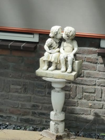 A stone statue of two children, a boy and a girl, is displayed outdoors in front of a brick wall. The children are depicted sitting next to each other and appear to be kissing. The girl is on the left, wearing a dress, and the boy is on the right, shirtless. The statue is mounted on a decorative pedestal. The background consists of a grey wall with a band of orange trim and a white window. The brick wall behind the statue is dark gray and weathered. The ground in front of the statue is paved with light-colored stones and scattered with dark gravel. This statue is located in Schaijk, Netherlands.