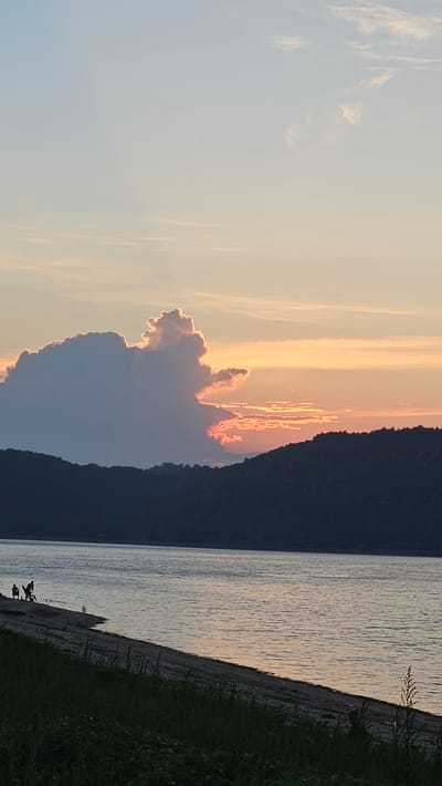 A serene sunset view captures the horizon over a wide body of water, likely a river or lake, in Kulishivka, Ukraine. Silhouetted against the dusky sky are rolling hills that form the distant shoreline. A striking cloud formation, illuminated by the setting sun with hues of orange and pink, dominates the sky above the hills. In the foreground, a narrow beach is visible, with a few individuals and possibly a dog casting long shadows as they stand near the water's edge. The water itself reflects the soft colors of the sky, creating a tranquil atmosphere. The lower edge of the image is lined with dark green vegetation.