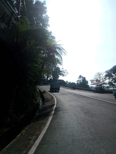 The image captures a winding asphalt road on the outskirts of Phouviang-Gnai, Laos. Lush, dense greenery cloaks the hillside to the left, juxtaposed against a bright, slightly hazy sky. A small, white vehicle travels up the road away from the viewer. Further up the road, a stationary food stand with a striped umbrella stands on the roadside with people around it. A portion of a silver car is visible on the right. The road is bordered by a white line.