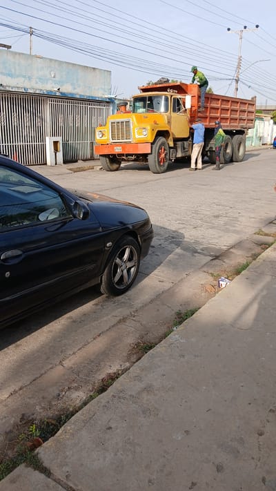 This image captures a daytime street scene in what appears to be an urban or suburban area, possibly Barcelona, Venezuela, under a clear, bright sky.

The central subjects are a yellow and orange dump truck with a rust-colored bed, and two men working around it. One man, dressed in a green long-sleeved shirt and a cap, stands on the side of the dump truck's bed, seemingly inspecting its contents or performing a task. Another man, wearing a blue shirt and light-colored pants, leans against the side of the truck's cab, appearing engaged in work near the front wheel. The truck's front bumper prominently displays a plate with the text "XV640".

To the right of the dump truck, further down the concrete street, a black sedan is parked. Its front passenger side, including a wheel with silver multi-spoke rims, is visible. The street surface is concrete, showing some signs of wear, and a concrete sidewalk runs along its right edge, with grass and weeds growing in its cracks.

In the background, behind the truck, there's a metal gate belonging to a property and a building featuring a weathered light-blue wall. Multiple utility lines are strung across the clear sky. The bright sunlight creates clear shadows on the ground, suggesting a sunny day. The scene depicts a moment of industrious activity related to the dump truck.