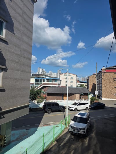 The image depicts a street scene in Jincheon-eup, South Korea, under a bright blue sky with scattered white clouds. The perspective is from a slightly elevated position, looking down at a street lined with buildings of varying architectural styles.
Several cars are parked along the street. A white SUV with a sunroof is prominently featured in the foreground, positioned on the right side of the road. Next to it, a dark-colored SUV is parked further down. Further up the street, another dark-colored car is visible. The street appears to be a typical urban or suburban thoroughfare.
The buildings showcase a mix of designs. Some are constructed with brick, while others have more modern facades with large windows. In the background, taller, multi-story buildings suggest a more densely populated area beyond the immediate street. There are also glimpses of greenery, including trees and bushes, interspersed with the structures.
A green metal fence runs along a portion of the street, separating it from what appears to be a paved area with a light blue surface, possibly a walkway or playground. Shadows are cast by the buildings and cars, indicating it is daytime, likely late morning or early afternoon due to the angle of the sun.
A few lines, likely power or communication cables, crisscross the sky. No people are visible in the image. The overall impression is a peaceful, everyday scene in a Korean town. License plate text "650 9053" is visible on the white SUV.