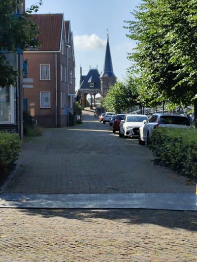 The image depicts a street in Sneek, Netherlands, featuring a row of parked cars on the right side of the road. The street is paved with cobblestones and lined with buildings on the left and trees on the right. In the background, a church tower with a spire is visible, along with an arched structure. The sky is blue with scattered clouds. There are no people visible in the image.