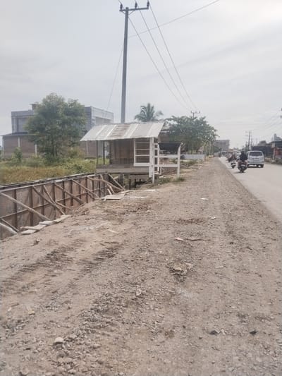 This daytime image captures a semi-urban or rural roadside scene in Barokulamgajah, Indonesia, under an overcast or hazy sky. In the foreground, a wide, unpaved dirt and gravel path stretches from the bottom left to the mid-right. To the left of this path, a long, deep ditch or canal is visible, lined with what appears to be wooden formwork and metal panels, supported by numerous wooden struts, suggesting ongoing construction or infrastructure work.

Adjacent to the ditch, a small, open-sided structure resembling a shack or stall stands, featuring a corrugated metal roof and white wooden framing. Further along, to the right of the dirt path, a paved road extends into the distance. Traffic is present on this road, including several motorbikes with riders (some wearing helmets) and a white minivan.

In the midground and background, a mix of buildings with simple architecture and various trees with green foliage line the road. Tall utility poles with multiple overhead power lines are prominent features against the light sky, indicating an established electrical infrastructure. No legible text is discernible on any objects, signs, or vehicles in the image. The scene depicts a functional but somewhat rustic environment with everyday activity.