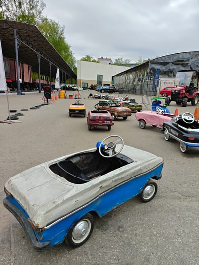 The photograph depicts an outdoor area in Arkhangel’skoye, Russia, showcasing a collection of vintage pedal cars. The cars are arranged haphazardly on a concrete surface, seemingly as a display or attraction. In the foreground, a blue and white pedal car with visible wear and tear is prominently featured. In the background, various other pedal cars in different colors and styles are visible, including a pink car, a black police car, a yellow car, and several others. A red tractor, along with a larger red train car behind a metal structure, is also visible in the background. A traffic light and safety cones indicate a possible makeshift traffic course or play area. The overall environment suggests an event or exhibition related to vintage vehicles. The sky is overcast, indicating cloudy weather conditions. Buildings and greenery can be seen in the distance.