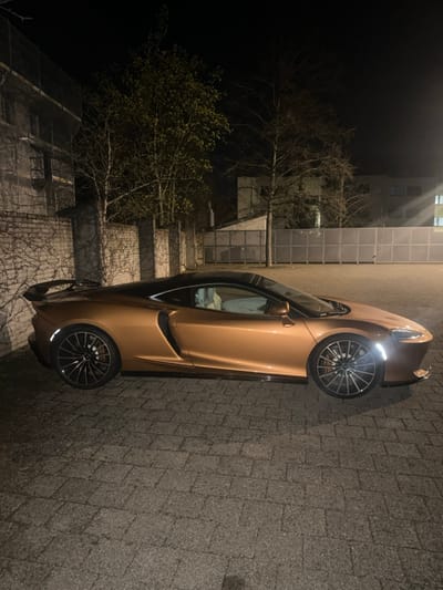 A metallic copper/bronze McLaren GT, featuring a black roof and light-colored interior, is parked facing right, displaying its left profile, on a paved surface composed of grey interlocking pavers. The front headlights of the vehicle are illuminated. Dark multi-spoke alloy wheels with orange/red brake calipers are visible. To the left of the car, a low brick wall, partially covered by climbing vegetation, runs parallel to the vehicle, with several deciduous trees and a building structure partially obscured by scaffolding beyond it. To the right, a tall, light-colored opaque barrier extends horizontally, behind which a building with illuminated windows and additional trees are discernible. The scene is illuminated by artificial light at night in Dietlikon, Switzerland. No individuals are present.