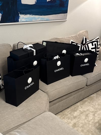The image shows a stack of six black Chanel shopping bags on a light-colored upholstered sofa with a geometric patterned pillow behind them. The sofa appears to be located indoors, possibly in a residential setting, as evidenced by the framed artwork hanging on a wall. While the city of Chula Vista, United States of America, is specified, there is no visual information within the image to confirm or establish this location. The focus is solely on the stacked shopping bags and the immediate surrounding objects.