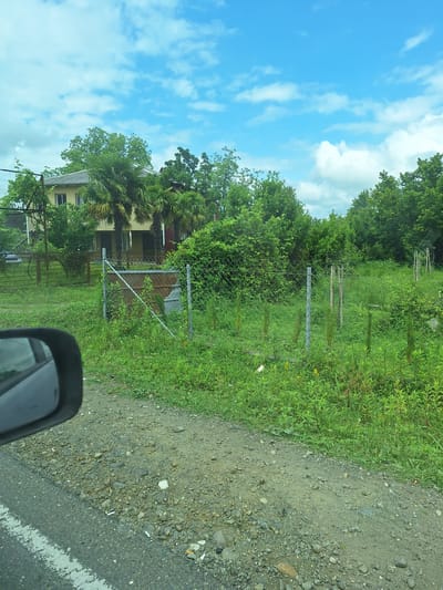 The image shows a roadside scene with a fence and a house in the background. The foreground consists of a gravel shoulder with some scattered rocks and debris, adjacent to an asphalt road with a white line. To the right of the road is a grassy area with tall weeds and a chain-link fence with metal posts. Behind the fence, there is a house with a yellow exterior and a tiled roof, partially obscured by lush green trees, including palm trees. The sky is blue with scattered white clouds. A car's side mirror is visible in the lower left corner of the image. The location is identified as ნიგვზიანის თემი, Georgia.