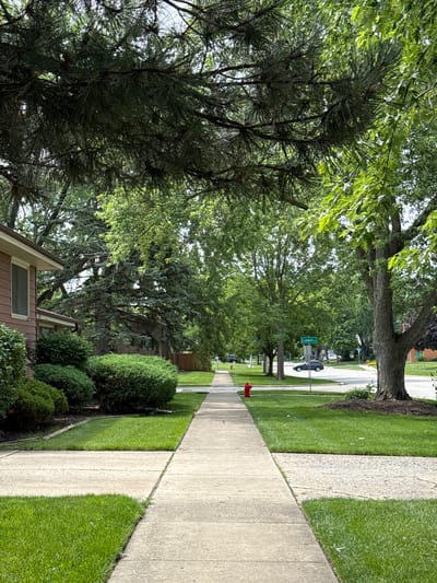 The image depicts a residential street in Woodridge, USA, on a bright, sunny day. A concrete sidewalk runs along the right side of the frame, bordered by a lush green lawn. The sidewalk leads into the distance, where it meets a street with cars parked and driving. Mature trees with full green foliage line the street, casting shadows on the sidewalk and lawns. A red fire hydrant is visible along the sidewalk. The scene suggests a quiet, suburban neighborhood. Text is visible on a green street sign that reads "Luxem." The overall mood is peaceful and serene, indicative of a typical summer day in the suburbs.