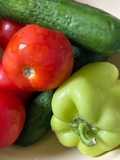 A close-up, top-down shot shows a collection of fresh, wet vegetables. In the foreground, a bright green bell pepper with a slightly wrinkled texture is prominent on the right. To its left and slightly behind, a ripe red tomato is visible, showing a slightly indented stem area. Another red tomato is partially visible in the lower left. Behind these, several dark green cucumbers with a textured surface fill the background. The vegetables appear to have been recently washed, as small water droplets are scattered across their surfaces. The scene is set against a light, neutral-colored background, likely a bowl or surface. No people or actions are depicted. The image is a still life of produce. The location of Mary City, Turkmenistan is not identifiable from the provided image.