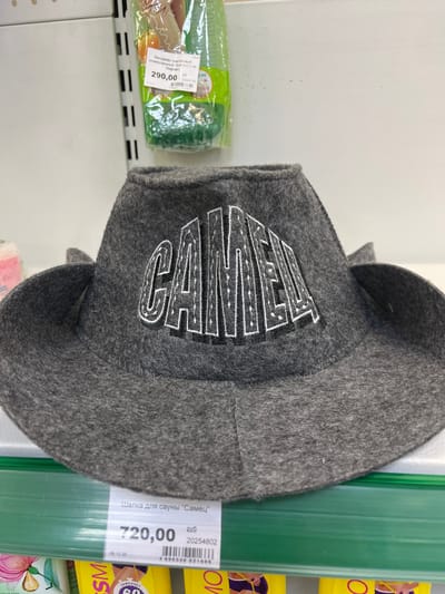 A grey felt hat with the word "САМЕЦ" embroidered in white and black lettering is displayed on a shelf in a store. The hat is positioned centrally in the frame, with its brim spread wide. Below the hat, a price tag is visible, reading "720,00 руб" and a barcode. To the left of the hat, a green shelf is visible, and on the far left of the image, a pink object with a floral design is partially visible. To the right of the hat, the background is a plain white wall. The image appears to be taken in a retail environment.
