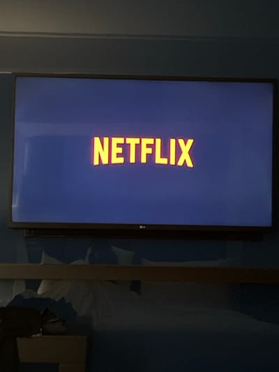 A flat-screen television is centrally positioned against a blue wall in a dimly lit indoor environment, identified as Bağcılar, Turkey. The television displays the bright yellow/orange "NETFLIX" logo on a dark blue background, indicating the platform is active. A faint LG logo is visible on the bottom bezel of the television. Below the television, a horizontal light brown wooden structure, possibly a shelf or a bed headboard, spans the width of the frame. Beneath this structure, a blue wall is visible, with white fabric (likely bedding) crumpled against it. A dark object, possibly a bag or folded clothing, rests on a lighter wooden surface at the bottom left, which appears to be part of a bedside table or bed frame. No individuals are present in the frame. Reflections are observable on the television screen, particularly on the left side.