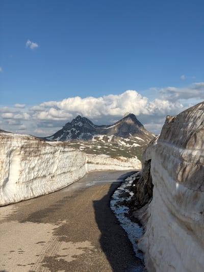 The image shows a mountain landscape under a partly cloudy, blue sky. In the center-left of the frame, a gray mountain peak is visible amidst white clouds. The foreground features a textured surface, with alternating bands of white and brown, possibly representing snow and gravel or dirt. To the right, a darker, uneven surface suggests a road or pathway, with visible tire tracks. The photograph appears to be taken at a high altitude, potentially in a mountainous region, but there is no evidence suggesting the city of Dubrovsko, Montenegro.