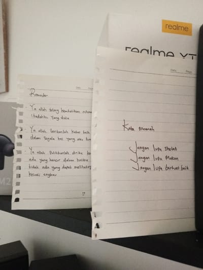The scene depicts an indoor environment in Al Haouafate, Morocco, featuring two handwritten notes. Two white, lined sheets of notebook paper, both exhibiting torn left edges consistent with removal from a binding, are positioned vertically against a light background.

The left paper, titled "Reminder" in cursive, contains three paragraphs of handwritten text in black ink, primarily in Indonesian, comprising a personal prayer or reflection. A small, hand-drawn smiley face is present at the bottom center of this note.

The right paper, partially overlapping the left, is titled "Kata Mamah" (Mom's words) in cursive. It lists three distinct pieces of advice or instructions handwritten in black ink, also in Indonesian. These instructions translate to "Don't forget to pray," "Don't forget to eat," and "Don't forget to do good."

Behind the papers, a white surface is visible, partially obscured by the notes. In the top right quadrant, a yellow rectangular sticker with the word "realme" in black font is visible, along with the partial text "realme YT" below it, indicating a product package. The papers rest on a dark surface, part of which exhibits a diagonal white stripe pattern. A dark, angular object is partially visible in the bottom-left corner of the frame. No individuals are present in the image. The notes suggest a personal space and serve as visual reminders or messages.