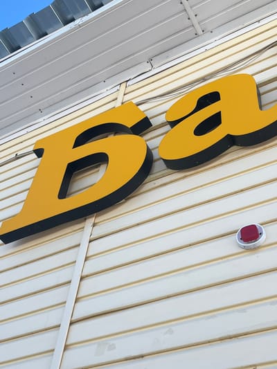 The image displays a close-up, upward-angled view of the exterior of a building located in Сундур, Russia. Large, three-dimensional yellow letters with black sides, spelling "Ба", are affixed to light-colored, horizontally paneled siding. The siding exhibits minor discoloration and wear, with visible fasteners. Above the letters, thin gray wires run horizontally across the siding. To the upper left, a section of a corrugated metal roof is visible against a clear blue sky. A small, circular white fixture housing a red rectangular light or sensor is mounted on the siding below and to the right of the yellow letters. No individuals or discernible activities are present in the scene.
