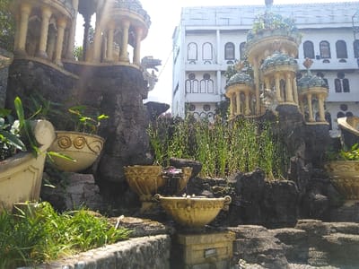 The image displays an elaborate ornamental garden feature located in Nová Bystřice, Czechia. The foreground and mid-ground are dominated by dark, textured artificial rock formations that serve as a base for multiple integrated decorative elements. These include several light-colored, classical-style miniature architectural structures featuring columns and domed roofs; some domes exhibit a teal coloration. One prominent structure on the left side includes multiple columns and a single dome, while a cluster of three smaller, similarly styled structures is visible toward the right.
Integrated into the rockwork are several ornate, beige-colored planters and bowls. A functional water feature is evident, with water cascading from an upper ornate bowl into a lower matching bowl. Vegetation is interspersed throughout the rock garden: tall, slender green plants, consistent with reeds or aquatic grasses, grow densely in various sections, particularly in the mid-ground. Broader-leafed green plants are present in some of the ornate planters on the left, and short green grass lines the bottom edge of the scene. A decorative element resembling a mythical creature's head is visible emerging from the rock formation on the left side, beneath one of the larger miniature structures.
In the background, a large, multi-story building with a light-colored facade (white or very light grey) is visible. The building features numerous windows, including both arched and rectangular designs, and exhibits decorative architectural details such as cornices or parapets along its roofline. The scene is illuminated by bright daylight, casting some areas into direct sunlight and creating strong shadows on the rock formations. No individuals are present in the image.