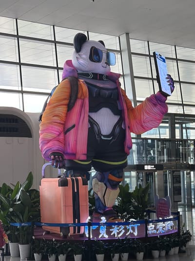This image depicts a large, stylized, anthropomorphic panda statue located in an airport terminal in Jianyang City, China. No human figures are present.

The central subject is a colossal panda figure, standing on a dark, illuminated base. The panda's head features a white and gray striped pattern, with black ears and prominent, square-rimmed blue sunglasses. It wears a futuristic, puffy jacket that transitions in color from orange on the right sleeve to vibrant pink and purple on the body and left sleeve, with black striped accents. A black backpack is visible on its back. The torso beneath the jacket appears to be armored with black and white plating, cinched by a thin yellow-green belt. The panda's legs are designed with a robotic or armored aesthetic, featuring segments of blue, orange, and white.

In its right hand (viewer's left), the panda holds the extendable handle of a peach-colored suitcase adorned with two vertical black straps. Its left hand (viewer's right) holds a large, illuminated smartphone vertically, displaying a map application with visible roads and some Chinese characters at the top, consistent with navigation software.

The base of the display features a blue rope barrier and is surrounded by numerous potted green plants, including peace lilies (Spathiphyllum) with white flowers, placed in white cylindrical pots. In front of the panda, an illuminated sign on the base displays Chinese characters, which translate to "National Treasure Lanterns Light Up..." (国宝彩灯 点亮...).

The environment suggests an airport terminal, characterized by a white paneled ceiling with recessed lighting. Large, multi-paned glass windows with dark frames form the background, revealing an overcast sky. The floor is a light-colored, reflective surface. In the mid-ground, a sign above glass doors clearly indicates "电梯 Elevator," pointing to an elevator bank. A large, curved white structure with a vent is visible to the left of the statue.