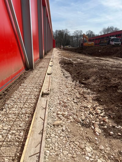 The image captures an active construction site in Donchevo, Bulgaria, under a partly cloudy sky. A large, red metal-clad building with prominent vertical grey structural support beams dominates the left side of the frame. Along the base of this structure, a prepared foundation trench contains a grid of rusted steel rebar laid over a coarse bed of light-colored gravel. Wooden formwork planks, some with exposed nails, border this rebar-and-gravel section. To the immediate right of the foundation work, the terrain transitions to dark brown, excavated soil and mud, displaying distinct vehicle tire tracks. In the mid-distance, an orange Hyundai excavator is positioned on a mound of earth, its boom raised. Adjacent to the excavator, a white Iveco dump truck is parked, facing left. A solitary individual, dressed in light-colored attire, is visible in the background near the center-left, facing away from the camera. A line of bare deciduous trees forms the distant backdrop beyond the construction zone.