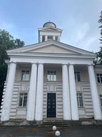 The image shows a close-up, low-angle view of the facade of a light-colored building with classical architectural elements, possibly a public building or historical site, located in Zavolzhye, Russia. The building features a prominent portico with six large white Ionic columns supporting a triangular pediment. Above the pediment, a dome-like structure topped with a spire is visible. The facade is decorated with decorative rectangular panels. There are multiple windows on the building, some with decorative metal grates. A dark double door is centered at the base of the portico. The ground in front of the building is paved and shows scattered fallen leaves. Two white spherical bollards are present in the foreground. The sky is overcast. The overall condition of the building appears somewhat weathered, with some cracks visible on the upper facade and evidence of wear on the stonework at the base. Trees with green foliage are visible on the sides of the building and partially obscure the upper structure. No people are visible in the image.
