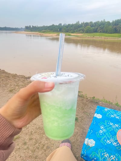 In Air Molek, Indonesia, a person's hand holds a clear plastic cup filled with a layered green and white beverage, topped with a blue and white striped straw. The cup's lid has some branding. The drink is positioned in the foreground, against a backdrop of a wide, muddy river and a lush green treeline. The ground in front of the river is sandy with patches of dry grass. To the right of the cup, a blue cloth with a white floral pattern is spread out, possibly as a makeshift tablecloth on a table. The sky is overcast and appears to be a hazy grey. The overall impression is of a casual outdoor setting by a river.