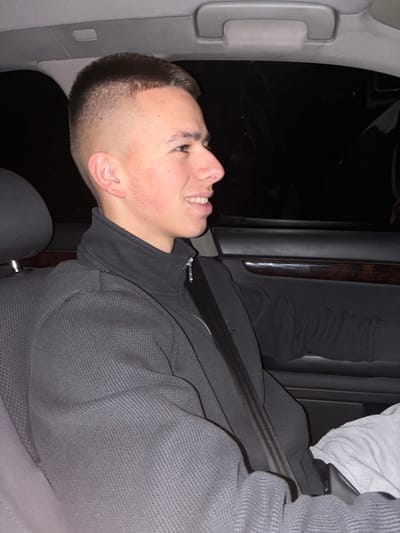 A young adult male is depicted seated in the front passenger seat of a vehicle in Zemen, Bulgaria. The subject, with a short fade haircut, light skin tone, and a small silver stud in his right nostril, is oriented in profile towards the right side of the frame, exhibiting a slight smile with teeth visible. He wears a dark grey waffle-knit texture zip-up jacket over a darker garment, with a silver zipper pull visible. A dark seatbelt with three parallel lighter stripes crosses his torso diagonally, buckling on his left side. Light-colored trousers are partially visible at the bottom right of the frame. The vehicle's interior features a dark grey or black door panel with a horizontal strip of wood-grain trim. A grey ceiling-mounted grab handle is visible above the dark side window, through which external darkness suggests nighttime conditions.