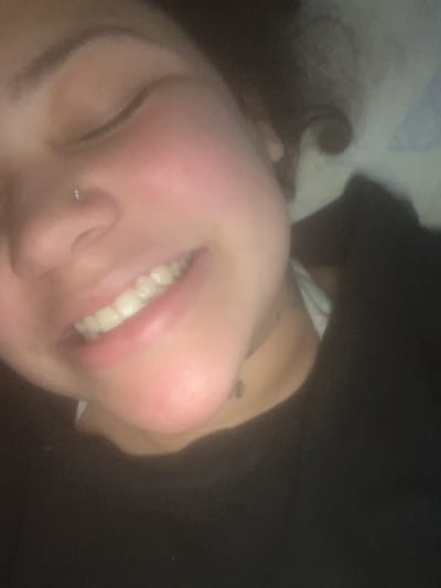 The scene is located in the city of Lancaster, United States of America. The image displays a close-up, slightly angled portrait of a single individual, appearing to be a young adult female, from the upper chest upwards. The individual's eyes are closed, and the mouth is open in a broad smile, revealing the upper row of teeth. A small, metallic stud piercing is present on the left nostril. The facial skin exhibits a reddish flush on the cheeks. The individual is wearing a dark, possibly black, garment over what appears to be a white undergarment, partially visible near the neck. A faint, dark mark, consistent with a small tattoo or shadow, is visible on the right side of the neck below the jawline. The background is indistinct and dark, with vague lighter shapes suggesting an indoor environment. Lighting appears soft and diffused, originating from the upper left.
