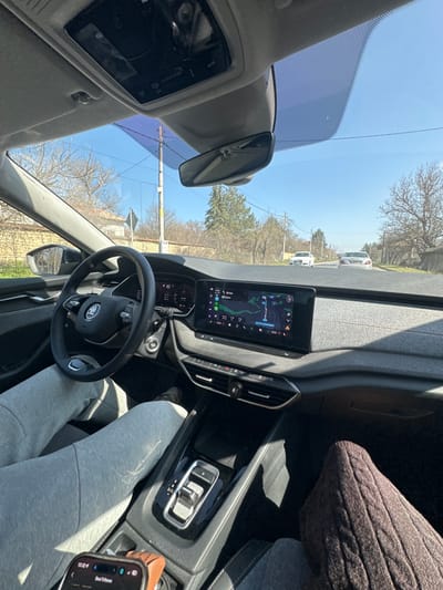 The image captures the interior of a modern passenger vehicle, likely a Skoda, from the front passenger's perspective, looking forward through the windshield onto a road in Ushintsi, Bulgaria, under a clear blue sky.

The driver's lower body is visible, wearing light grey sweatpants and light-colored shoes, with the left foot positioned on a pedal. The steering wheel, bearing a Skoda emblem, is turned slightly left. The digital instrument cluster displays a speed of "43" and an exterior temperature of "4.5°C". A large central infotainment screen shows a navigation map with a highlighted blue route and a current location marker. Below the screen are air vents and climate controls. The center console features an automatic gear selector. A smartphone in a brown case rests in a cup holder, displaying "13:32" and "Stael Trifonov". The front passenger's legs are partially visible, clad in dark brown knitted pants. An overhead console and rearview mirror are visible above the windshield.

Outside the vehicle, a two-lane asphalt road extends forward. In the distance, a white Audi sedan precedes a lighter-colored Mercedes-Benz sedan, both traveling in the same direction. On the left side of the road, a low stone wall is visible, accompanied by bare deciduous trees and a utility pole. A dark, rectangular road sign is also present on the left. On the right side, several bare deciduous trees and one taller evergreen tree line the road. The overall environment suggests a rural or semi-urban setting during late winter or early spring.