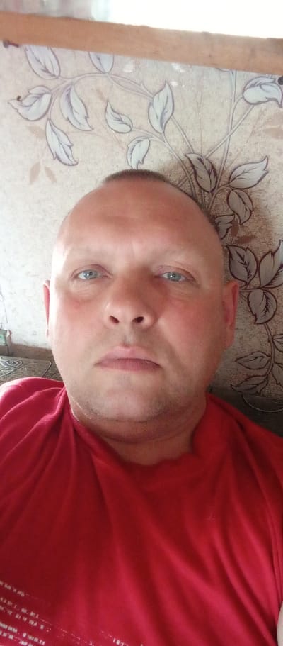This is a selfie of a middle-aged Caucasian man. He has a shaved head and light blue eyes. He is wearing a bright red t-shirt. The background appears to be indoors, with wallpaper featuring a floral or leafy pattern. The lighting suggests daytime. The man has a neutral expression. There is some visible stubble on his chin. The overall setting appears to be a casual indoor environment. There is no discernible text in the image.